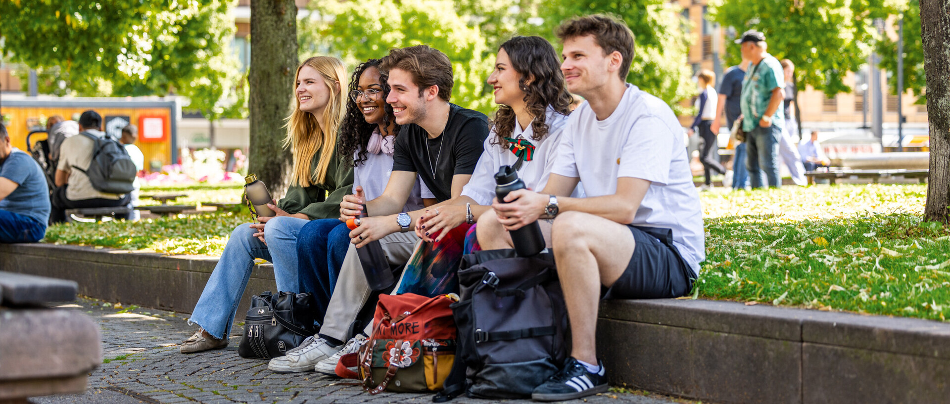 Eine Gruppe von fünf Studierenden sitzen an einem sonnigen Tag am Paradeplatz. Sie scheinen glücklich zu sein und gute Laune zu haben. Im Hintergrund sind viele andere Menschen zu sehen.