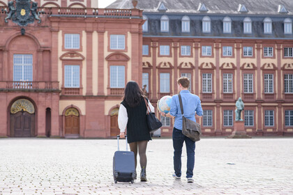 Zwei Personen laufen nebeneinander auf das Schloss der Uni Mannheim zu. Eine Person zieht einen schwarzen Koffer, während die andere Person eine Tasche sowien einen Globus trägt.