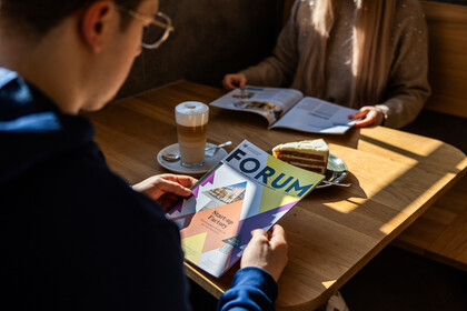 Zwei Personen lesen das Uni-Magazin „FORUM“ bei Kaffee und Kuchen in einem Café.
