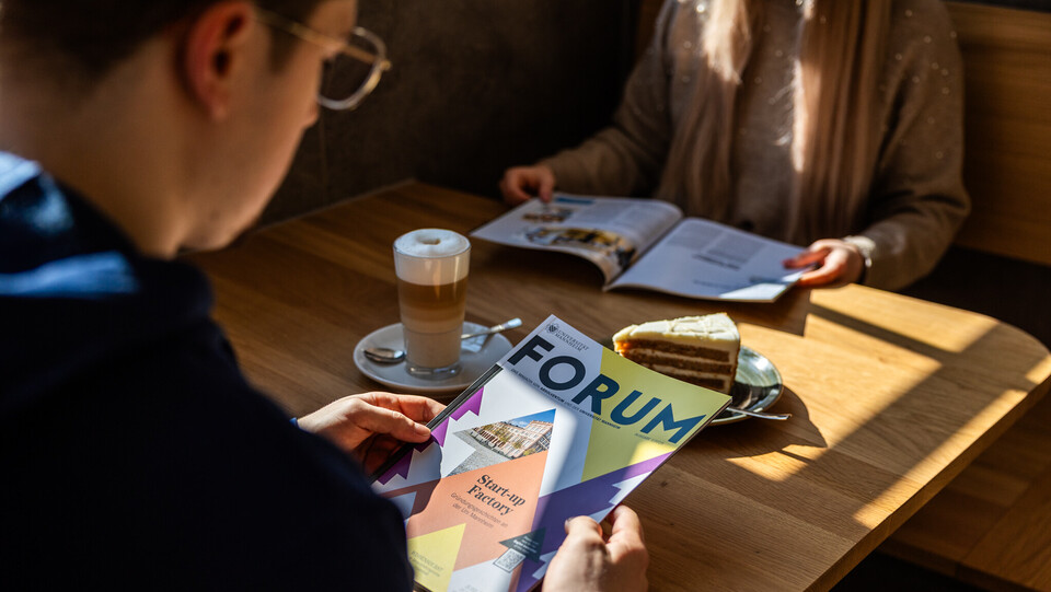 Zwei Personen lesen das Uni-Magazin „FORUM“ bei Kaffee und Kuchen in einem Café.