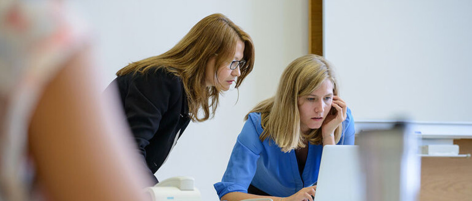 Zwei Frauen schauen zusammen auf den Bildschirm eines Laptops. Sie stehen vorne an der Tafel.