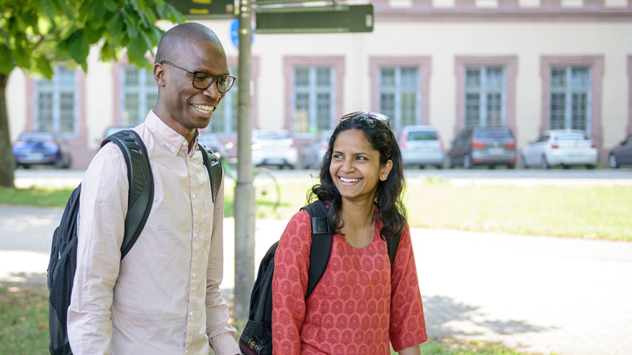 Zwei Personen laufen lachend über das Campus-Gelände. Sie tragen beide schwarze Rucksäcke.