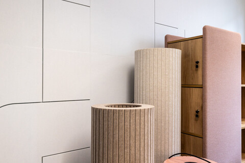 Auf dem Foto sind Details einer weißen schallhemmenden Fließwand zu sehen. Davor stehen stoffbezogene Säulen. Man sieht die Hälfte eines Raumteilers.