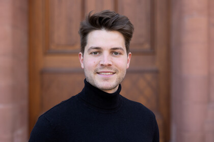 Felix Rusche. He is wearing a black sweater and is standing in front of a brown door.