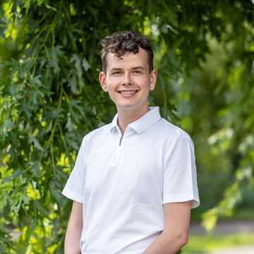 Felix Haas steht im weißen Kurzarmhemd vor einem grünen Hintergrund mit Bäumen. Er lächelt in die Kamera.