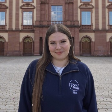 Martina has long, light brown hair and is wearing a dark blue pullover of the departmental student committee. She is standing in front of the Schloss and smiling at the camera.