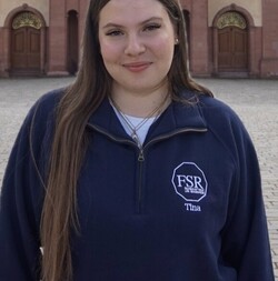 Martina mit langen, hellbraunen Haaren trägt einen dunkelblauen Pullover der Fachschaft und lächelt freundlich in die Kamera vor dem Mannheimer Schloss