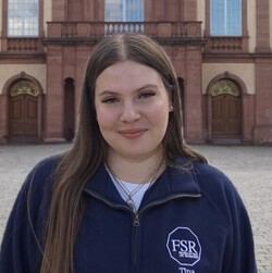 Martina has long, light brown hair and is wearing a dark blue pullover of the departmental student committee. She is standing in front of the Schloss and smiling at the camera.