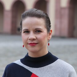 Daniele Hohmann hat ihre braunen Haare zusammengebunden. Sie trägt rote Hängeohrringe und einen Pullover, der rote, schwarze und graue Anteile hat. 