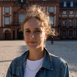 Ein Porträt von Valeria Pinto auf dem Ehrenhof vor dem Mannheimer Schloss. Sie trägt lockiges blondes Haar, ein weißes Oberteil und darüber eine blaue Jeansjacke.