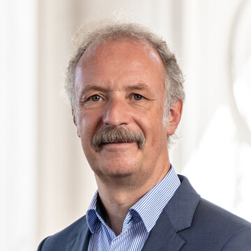 Prof. Dr. Moritz Fleischmann hat graue Haare und einen Schnauzer. Er trägt ein dunkles Jackett und ein gestreiftes Hemd.