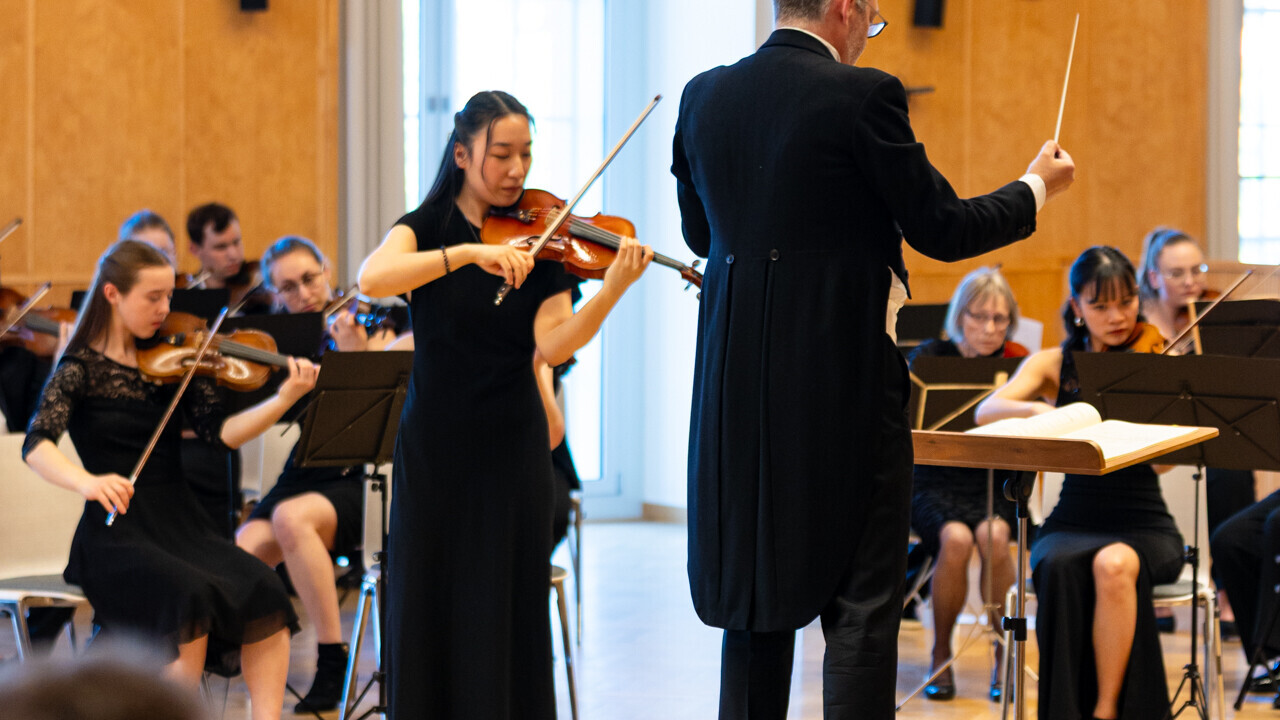 Auftritt des Uniorchesters. Dirigent steht vor den Studierenden, die jeweils ein Instrument spielen. Alle sind schwarz gekleidet.