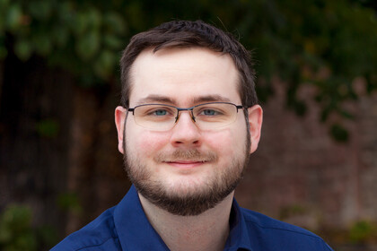 Tobias Rettig hat kurze dunkle Haare und einen Bart. Er trägt eine Brille und ein blaues Hemd.