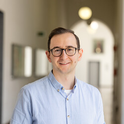 Can Yilanci hat kurze braune Haare. Er trägt eine Brille und ein hellblaues Hemd und steht im Universitätsflur.
