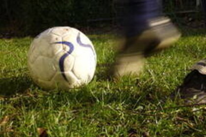 Ein weißer Fußball mit blauen Verzierungen liegt auf einem grünen Rasen und ist kurz davor, vor einem schwarzen Fußballschuh mit hoher Geschwindigkeit in die Ferne geschossen zu werden.