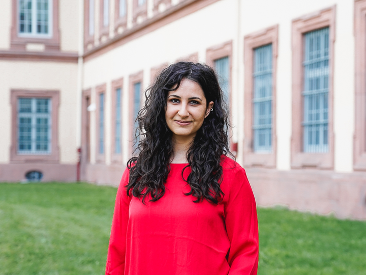 Abir Al-Laham hat lange lockige schwarze Haare. Sie trägt ein leuchtend rotes Oberteil und steht vor dem Schloss.