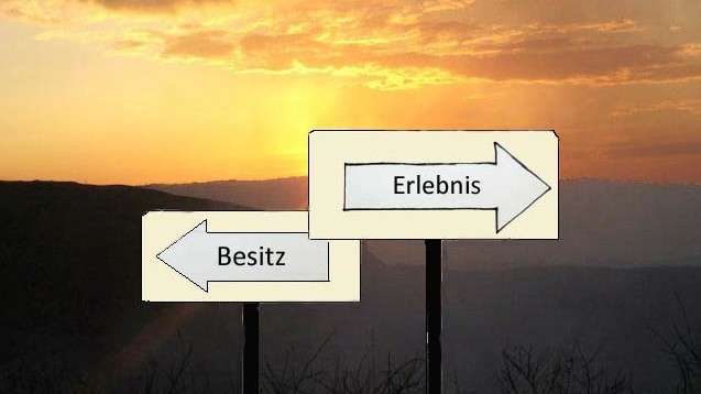 Vor dem Hintergund einer hügeligen Landschaft im Sonnenuntergang stehen zwei Wegweiser in entgegengesetze Richtungen, einer mit der Aufschrift "Besitz", ein anderer mit der Aufschrift "Erlebnis".