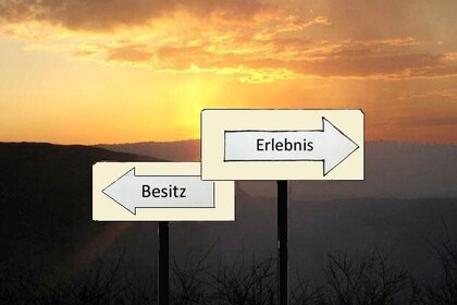 Vor dem Hintergund einer hügeligen Landschaft im Sonnenuntergang stehen zwei Wegweiser in entgegengesetze Richtungen, einer mit der Aufschrift "Besitz", ein anderer mit der Aufschrift "Erlebnis".