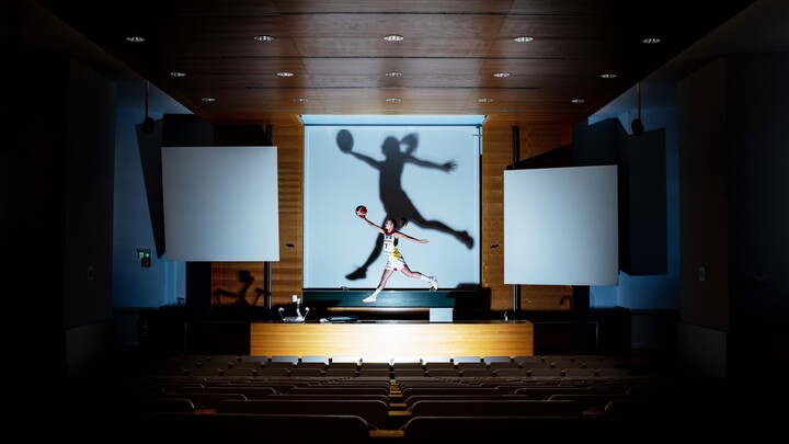 Eine Person trägt Sportkleidung und springt über einem Pult in einem leeren Hörsaal. In ihren rechten Hand hält sie ausgestreckt einen Handball.