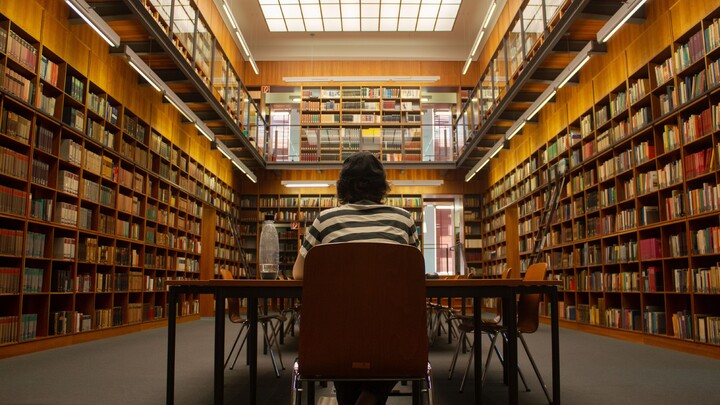 Wissen Eine studierende Person sitzt mit dem Rücken zur Kamera an einem Schreibtisch in einer Bibliothek.