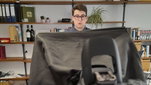 Eine Person steht vor einem Bücherregal und hinter einer Kamera. Die Kamera steht im Vordergrund und nimmt einen Großteil des Bildes ein.