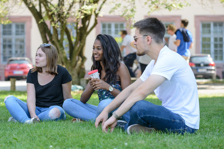 Zwei Studentinnen und ein Student sitzen auf einer Wiese und unterhalten sich. Sie tragen T-Shirts und Sonnenbrillen.
