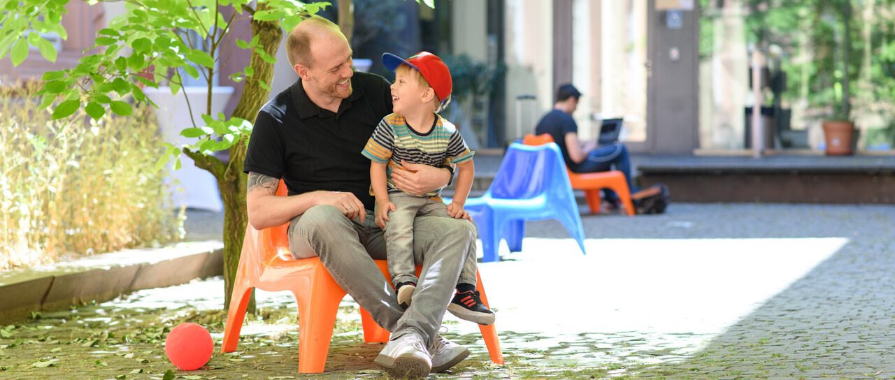 Father and son are sitting on the campus of the University of Mannheim and smiling at each other.