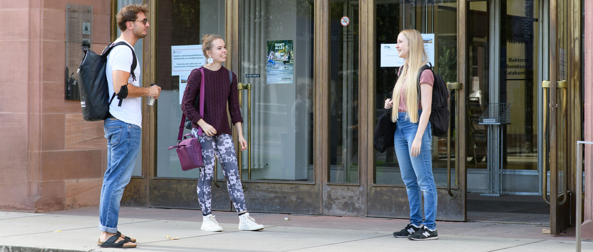Three students stand about 1.5 meters apart in front of the main entrance to the University of Mannheim and talk to each other.
