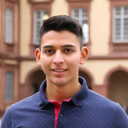Juan David hat kurze dunkelbraune Haare, trägt ein blaues Poloshirt und lächelt.