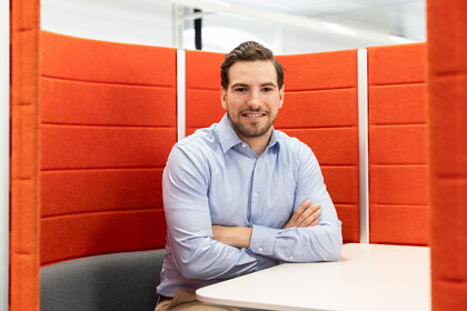 Porträt von Jan Sindelar. Er sitzt an einem Tisch, die Wand hinter ihm ist orange. Er hat kurze braune Haare, trägt ein hellblaues Hemd und schaut in die Kamera.