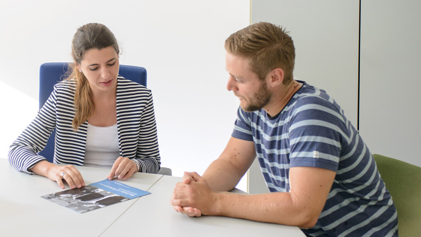 Eine Mitarbeiterin berät einen Studenten sitzend am Tisch mit einer Broschüre.