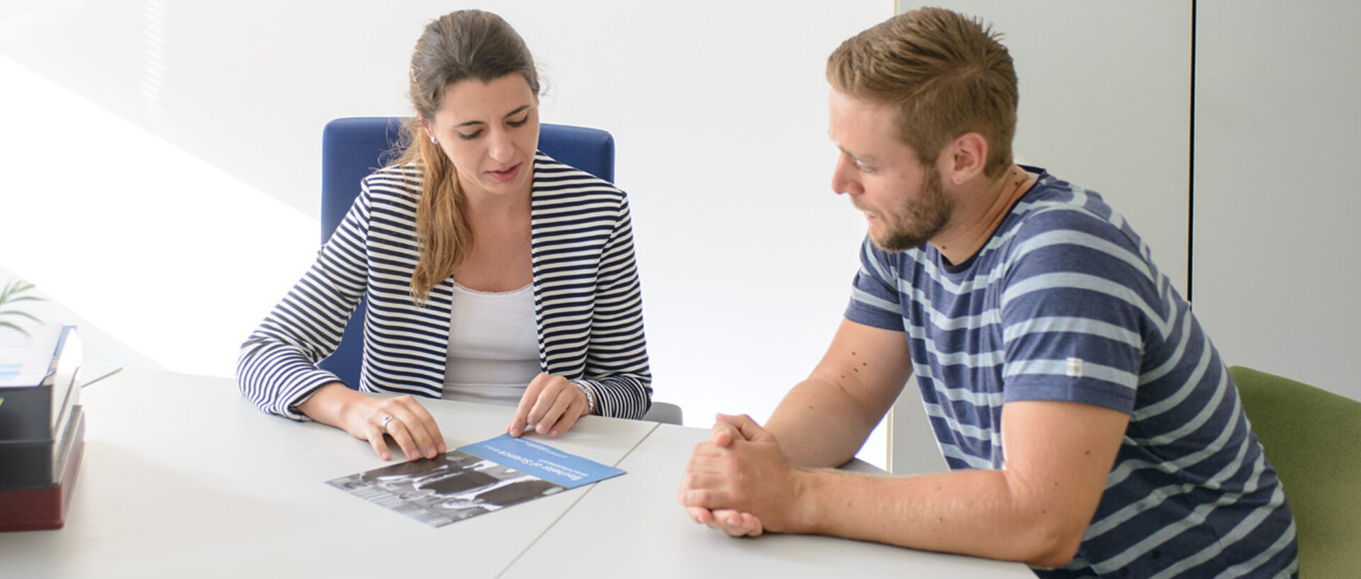 Eine Mitarbeiterin berät einen Studenten sitzend am Tisch mit einer Broschüre.