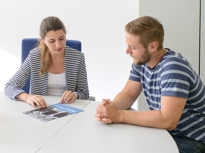 Eine Mitarbeiterin berät einen Studenten sitzend am Tisch mit einer Broschüre.