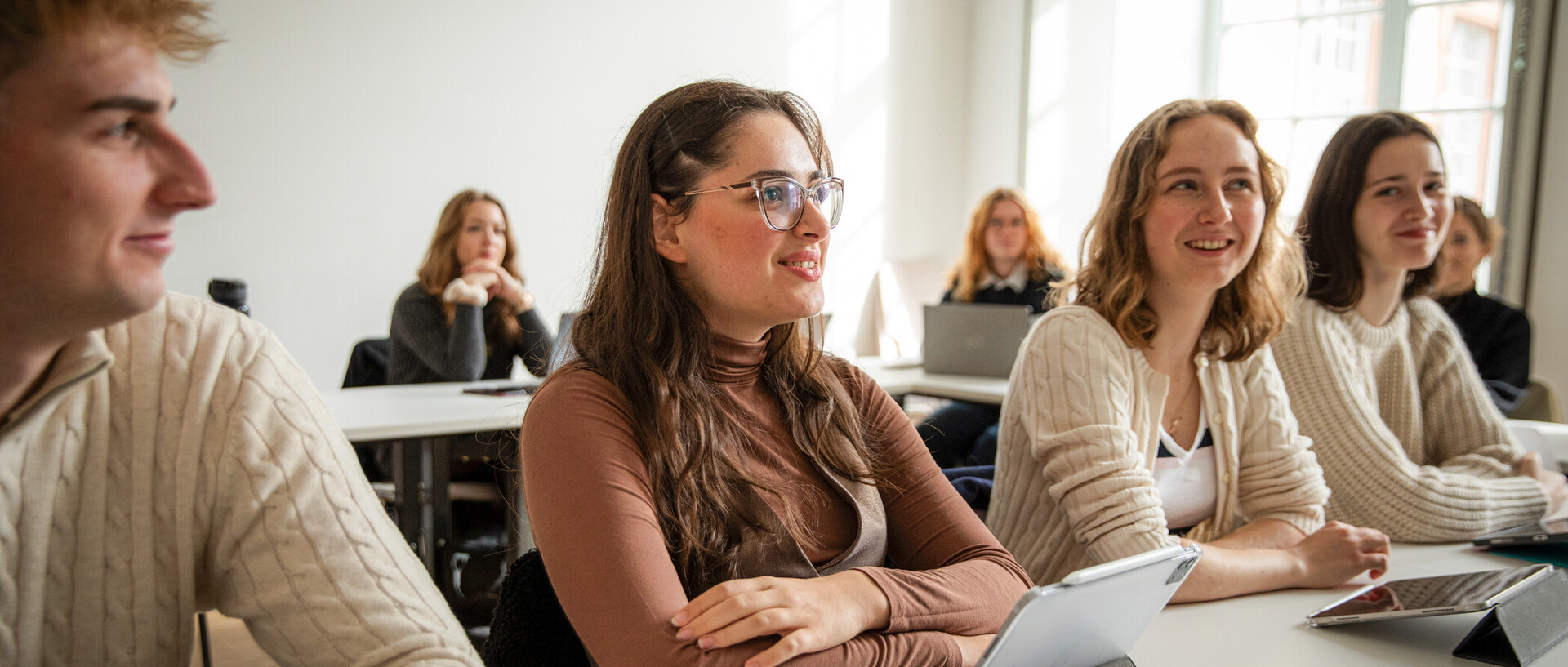 Studierende sitzen in einem Seminarraum an Tischen und schauen Richtung Tafel