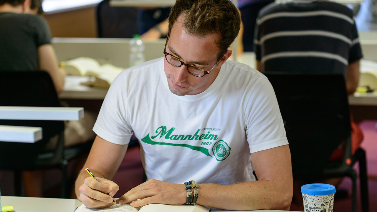 Ein Student liest in einem Buch. Er trägt eine Brille und ein weißes T-Shirt mit der Aufschrift "Universität Mannheim".