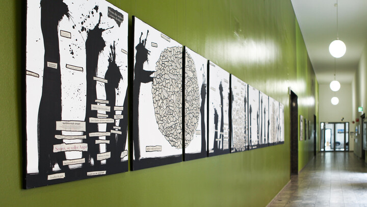 In einem Flur mit grüner Wand hängen Kunstwerke bestehend aus Zeitungscollagen und schwarzen Farbklecksen.