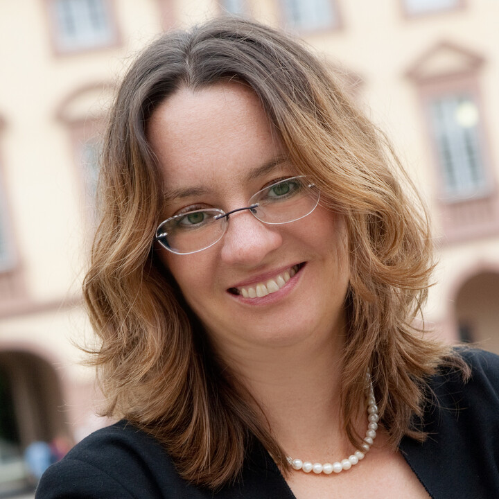 Prof. Michèle Tertilt, Ph.D. vor dem Schloss, sie hat schulterlange blond-braune gewellte Haare, eine ovale Brille, eine Perlenkette und einen schwarzen Anzug