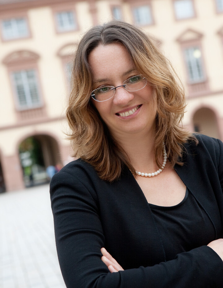 Prof. Michèle Tertilt, Ph.D. vor dem Schloss, sie hat schulterlange blond-braune gewellte Haare, eine ovale Brille, eine Perlenkette und einen schwarzen Anzug
