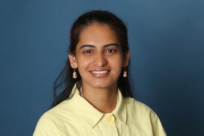 Portraitfoto von Pooja Rani. Sie trägt ein gelbes Oberteil und lächelt vor einem blauen Hintergrund in die Kamera.