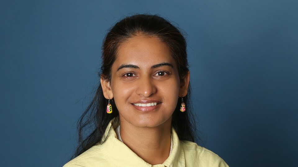 Portraitfoto von Pooja Rani. Sie trägt ein gelbes Oberteil und lächelt vor einem blauen Hintergrund in die Kamera.