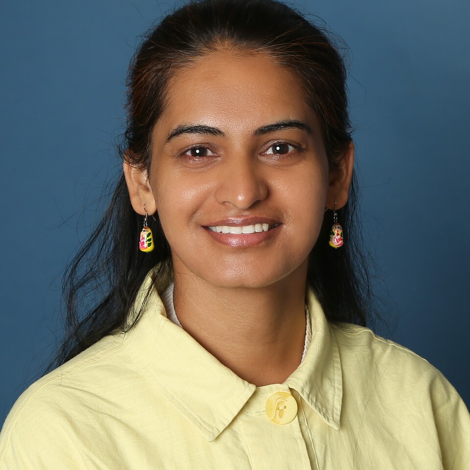 Portraitfoto von Pooja Rani. Sie trägt ein gelbes Oberteil und lächelt vor einem blauen Hintergrund in die Kamera.