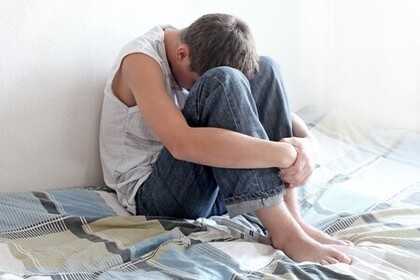 Ein Teenager sitzt mit angezogenen Beinen auf seinem Bett. Sein Kopf ist auf den Knien abgelegt und die Arme um seine Beine geschlungen.