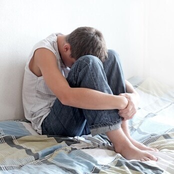 Ein Teenager sitzt mit angezogenen Beinen auf seinem Bett. Sein Kopf ist auf den Knien abgelegt und die Arme um seine Beine geschlungen.