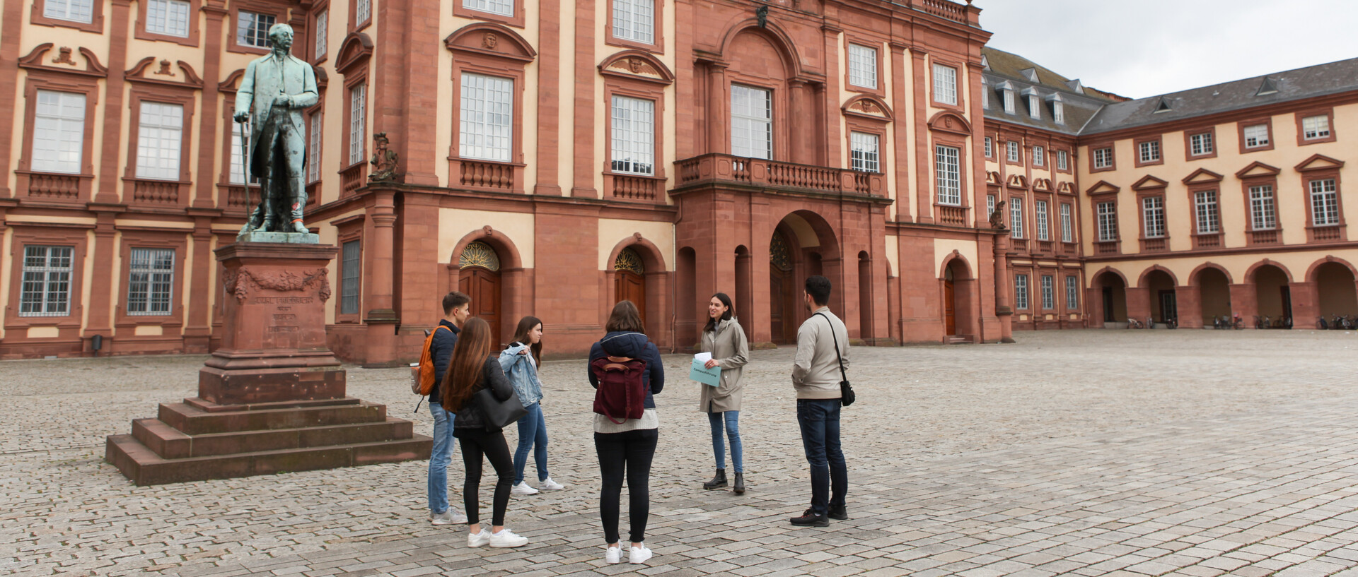 Vier Studierende hören einer Studentin beim Erzählen zu. Sie stehen auf dem Ehrenhof der Universität Mannheim. Im Hintergrund befindet sich das Mannheimer Schloss.