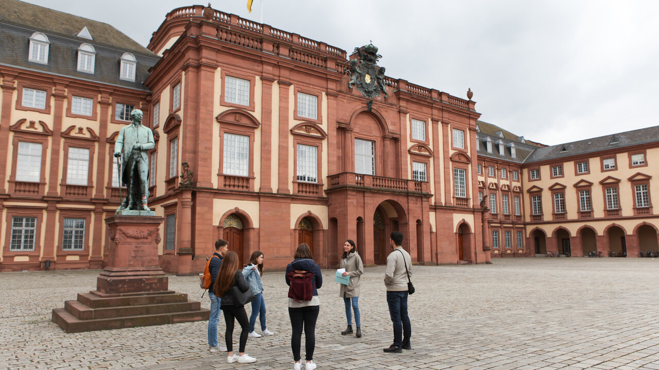 Vier Studierende hören einer Studentin beim Erzählen zu. Sie stehen auf dem Ehrenhof der Universität Mannheim. Im Hintergrund befindet sich das Mannheimer Schloss.