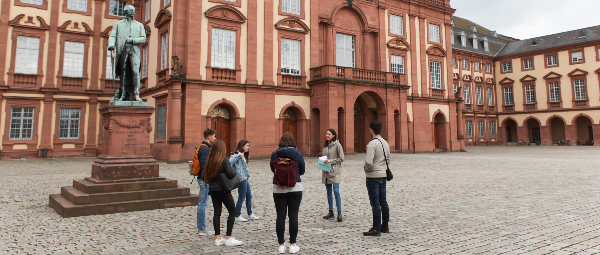 Vier Studierende hören einer Studentin beim Erzählen zu. Sie stehen auf dem Ehrenhof der Universität Mannheim. Im Hintergrund befindet sich das Mannheimer Schloss.