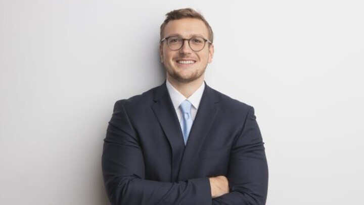 Pascal Schrader. He is wearing a black suit and is standing in front of a white wall.
