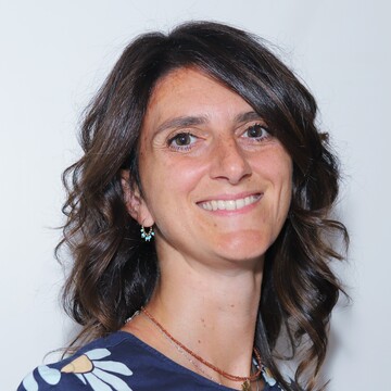 Mariateresa Cecalupo vor einem hellblauen Hintergrund. Sie hat schulterlanges, gelocktes Haar und trägt ein dunkelblaues Oberteil.