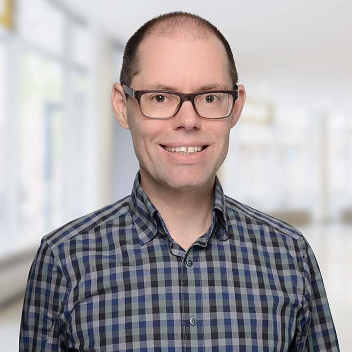 Portrait von Martin Peitz. Er trägt ein karriertes dunkelblau-schwarzes Hemd und lächelt.