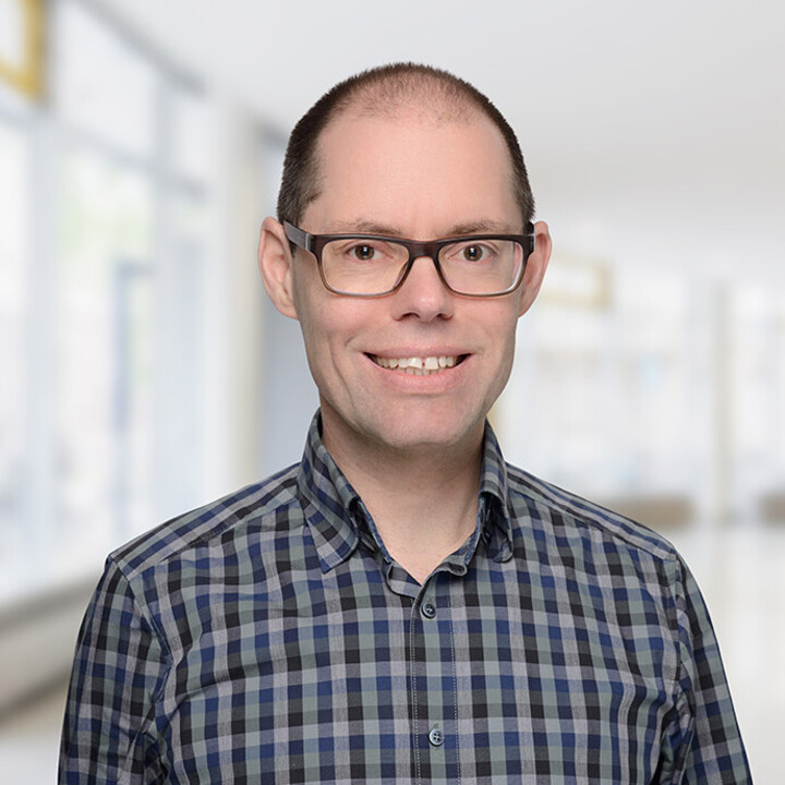 Portrait von Martin Peitz. Er trägt ein karriertes dunkelblau-schwarzes Hemd und lächelt.